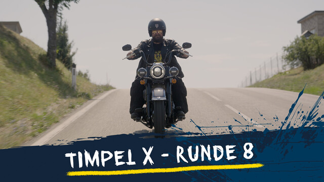 Timpel sitztauf einem Motorrad und fährt auf einer Freilandstraße, links und rechts sind Wiese und Bäume
