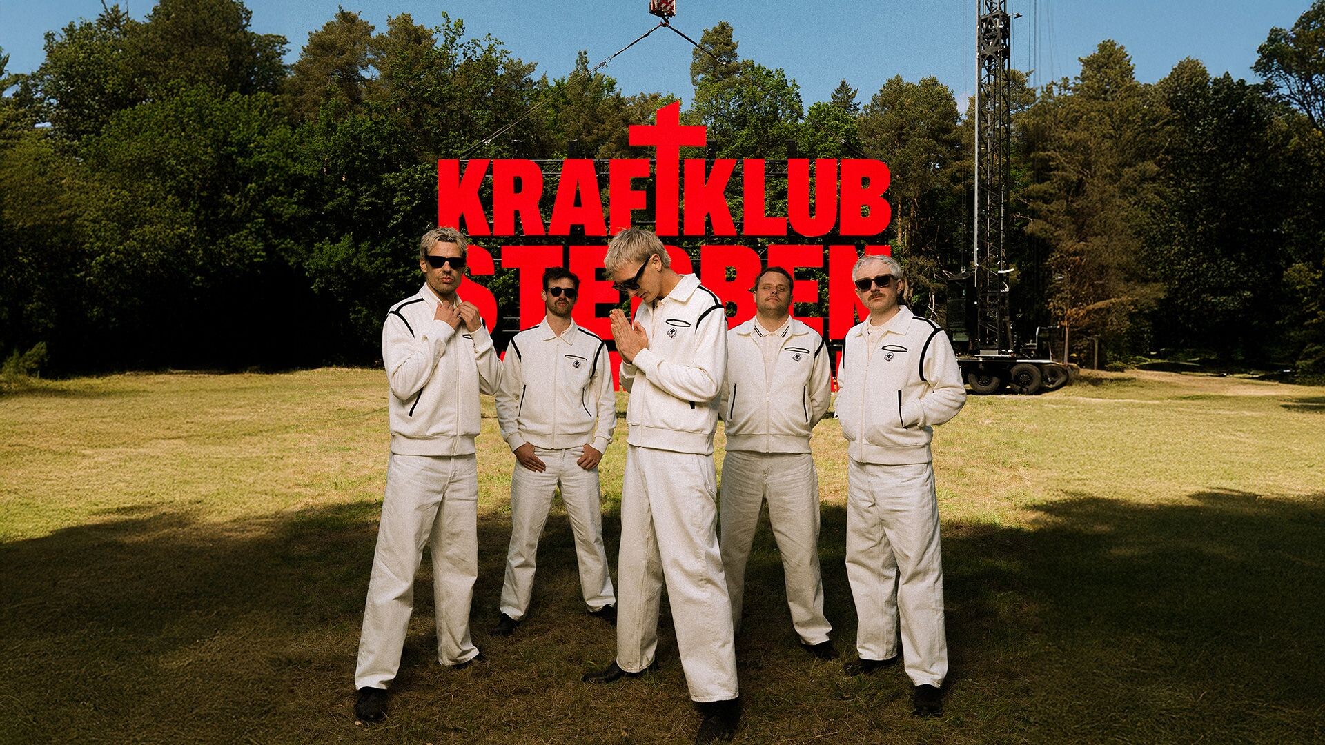 die Band steht auf einer Wiese, dahinter sind ein Wald und ein Kran zu sehen