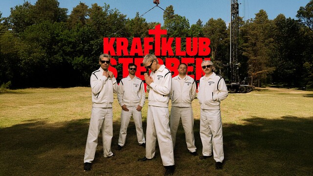 die Band steht auf einer Wiese, dahinter sind ein Wald und ein Kran zu sehen