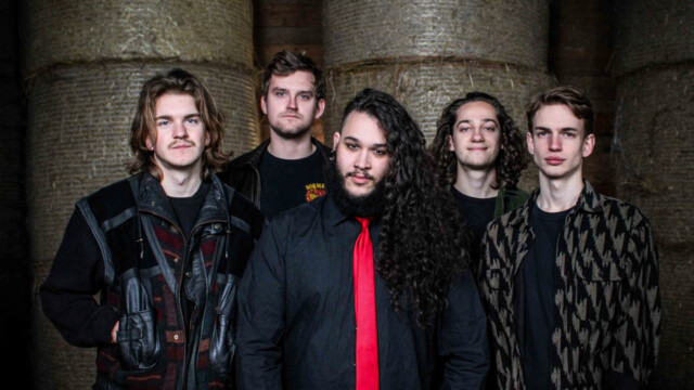 Die Band One Last Glance posieren in schwarzer beziehungsweise dunkel gehaltener Kleidung vor kaum Erkennbaren Heuballen hinter Ihenen. Der Sänger trägt eine rote Krawatte