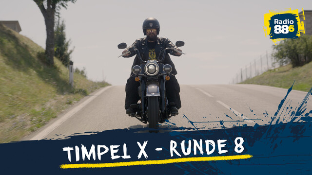 Timpel sitzt auf einem Motorrad und fährt auf einer Freilandstraße, links und rechts sind Wiese und Bäume