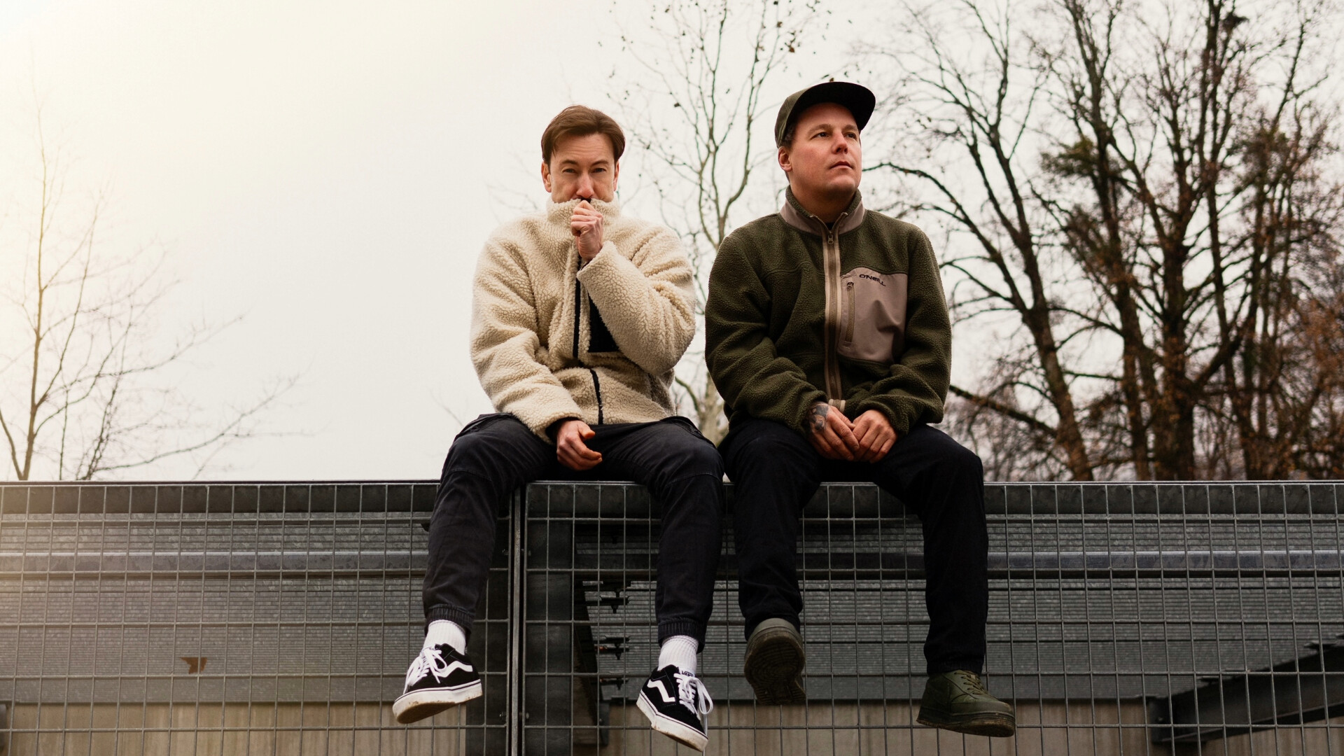 Coperniquo sitzen mit Herbstlicher Kleidung auf einem Metallzaun, Baumkronen und grauer Himmel im Hintergrund